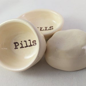 May include: Three small, white ceramic dishes with the word "Pills" written on one of them. The dishes are ideal for holding small items such as pills, jewellery, or other trinkets.