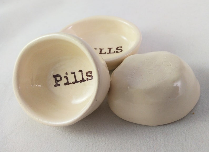 May include: Three small, white ceramic dishes with the word "Pills" written on one of them. The dishes are ideal for holding small items such as pills, jewellery, or other trinkets.