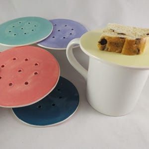May include: Four ceramic coasters in different colors: teal, lavender, coral, and navy blue. Each coaster has a circular shape with a white rim and multiple small holes. A white ceramic mug with a yellow saucer and a slice of bread on top.
