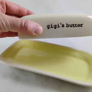 May include: Cream-coloured butter dish with a pale yellow interior. The lid is inscribed with "gigi's butter" in black lettering. The dish is rectangular with rounded edges.