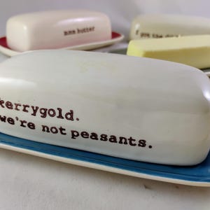 May include: White ceramic butter dish with the text "kerrygold. we're not peasants." on a blue rectangular plate. Other butter dishes are visible in the background, some with butter inside. The butter dishes are on a white surface.