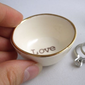 May include: A small white ceramic ring dish with a gold rim and the word "LOVE" inscribed on the inside. The dish is sitting on a white surface.