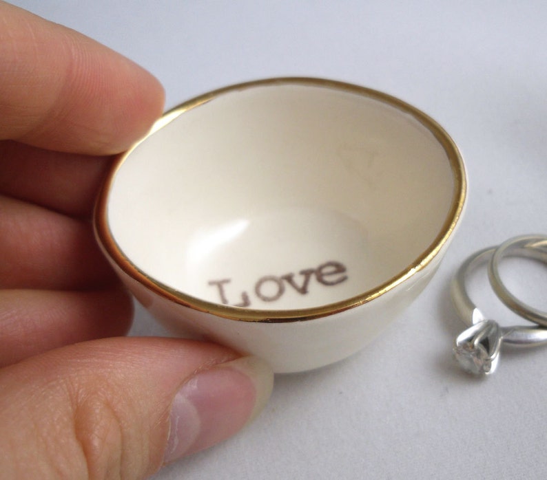 May include: A small white ceramic ring dish with a gold rim and the word "LOVE" inscribed on the inside. The dish is sitting on a white surface.