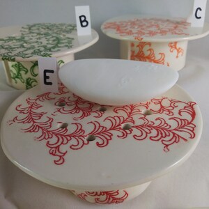 May include: Three ceramic soap dishes with different floral designs. The dish in the foreground has a red floral design and a white bar of soap on top. The dish in the background on the left has a green floral design. The dish in the background on the right has an orange floral design.