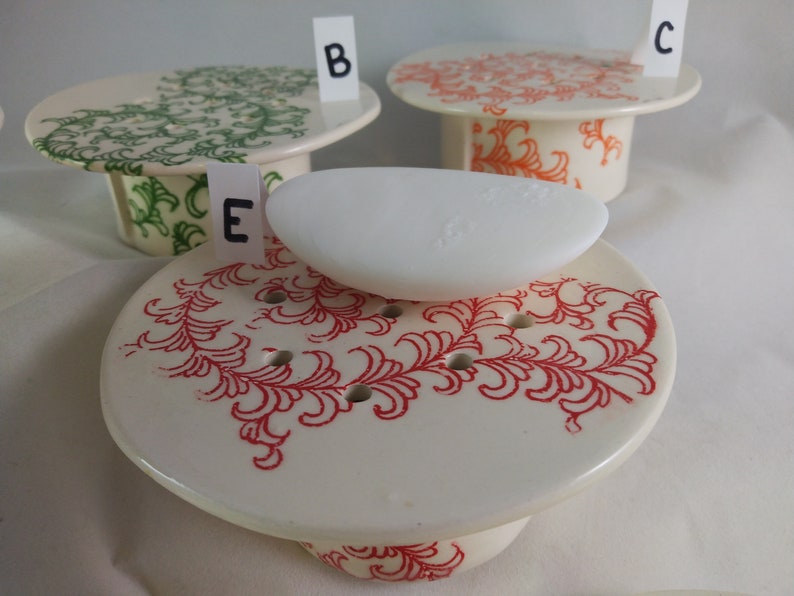 May include: Three ceramic soap dishes with different floral designs. The dish in the foreground has a red floral design and a white bar of soap on top. The dish in the background on the left has a green floral design. The dish in the background on the right has an orange floral design.
