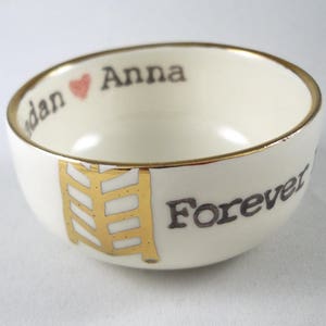May include: White ceramic bowl with a gold rim and a gold design. The bowl has the words "Anna" and "Forever" written on it in black.