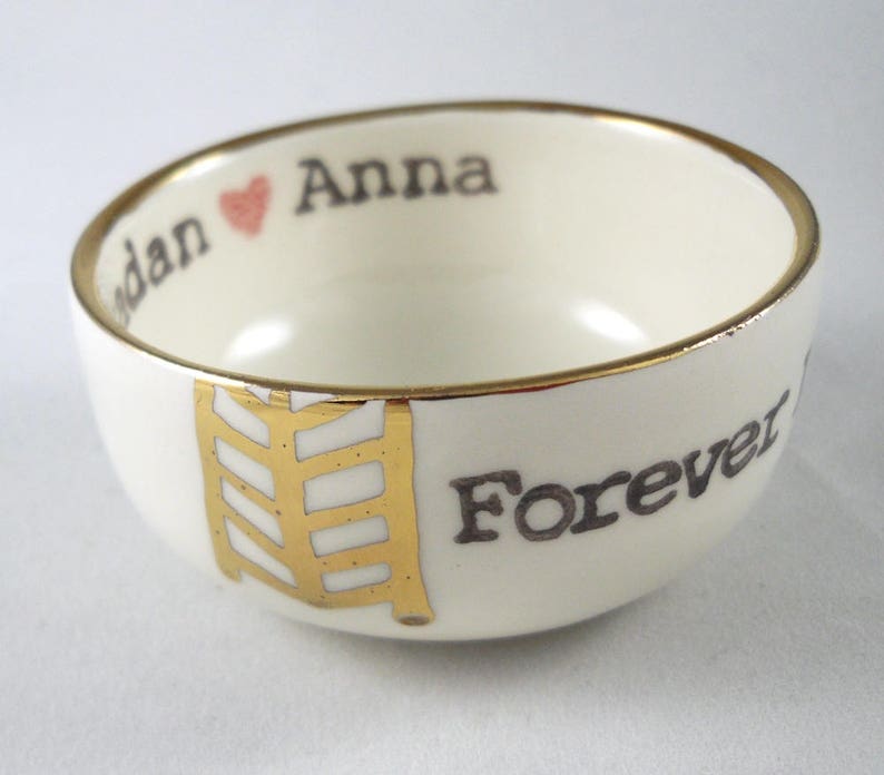 May include: White ceramic bowl with a gold rim and a gold design. The bowl has the words "Anna" and "Forever" written on it in black.
