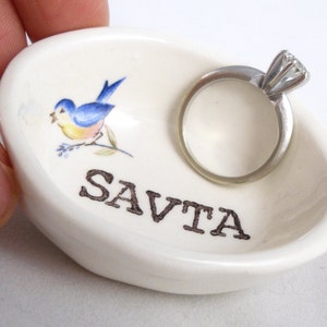 May include: White ceramic ring dish with a bluebird design and the word "SAVTA" printed on the bottom. A silver ring with a diamond is sitting in the dish.