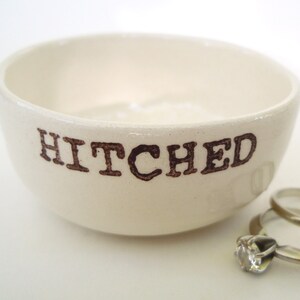May include: White ceramic ring dish with the word "HITCHED" printed in brown lettering. The dish is empty, but there are two gold wedding bands sitting next to it.
