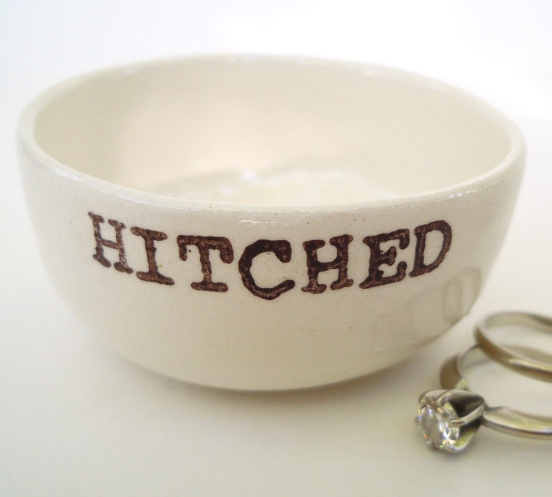 May include: White ceramic ring dish with the word "HITCHED" printed in brown lettering. The dish is empty, but there are two gold wedding bands sitting next to it.