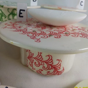 May include: White ceramic soap dish with a red floral design. The dish has a raised edge and a small, white soap bar on top. The dish is labeled with the letter "E".