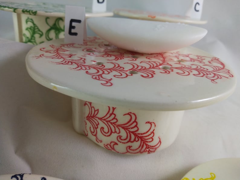 May include: White ceramic soap dish with a red floral design. The dish has a raised edge and a small, white soap bar on top. The dish is labeled with the letter "E".