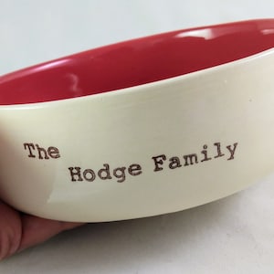Può includere: Una ciotola in ceramica bianca per animali domestici con interno rosso. La ciotola ha le parole "The Hodge Family" incise all'esterno.
