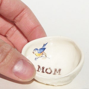 May include: White ceramic trinket dish with a blue bird and green leaves design. The word "MOM" is written in brown lettering on the bottom of the dish.