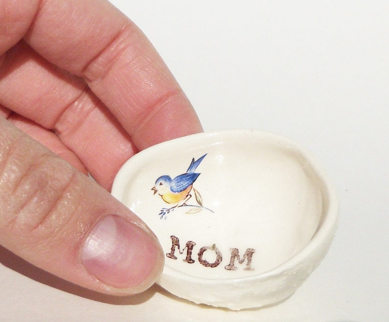 May include: White ceramic trinket dish with a blue bird and green leaves design. The word "MOM" is written in brown lettering on the bottom of the dish.