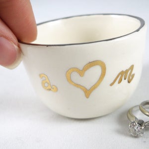 Puede incluir: Plato de cerámica blanco para anillos con un corazón dorado y las letras "a" y "m" en la parte delantera. Dos anillos de oro están sobre una superficie blanca junto al plato.