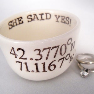 May include: Cream-colored ceramic ring dish with the text "SHE SAID YES!" and coordinates. Two silver rings, one with a diamond, are next to the dish. The dish is slightly tilted, showcasing the interior.