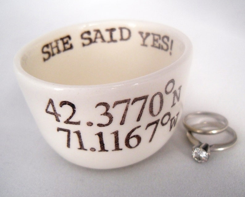 May include: Cream-colored ceramic ring dish with the text "SHE SAID YES!" and coordinates. Two silver rings, one with a diamond, are next to the dish. The dish is slightly tilted, showcasing the interior.