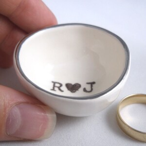 May include: White ceramic ring dish with a silver rim and the initials "R&J" with a heart in between.