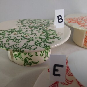 May include: Three ceramic cake stands with different designs. The first stand has a green floral pattern, the second stand has an orange floral pattern, and the third stand is plain white. The stands are labeled with the letters "B" and "E".