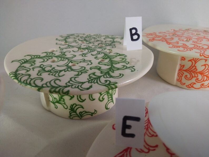 May include: Three ceramic cake stands with different designs. The first stand has a green floral pattern, the second stand has an orange floral pattern, and the third stand is plain white. The stands are labeled with the letters "B" and "E".