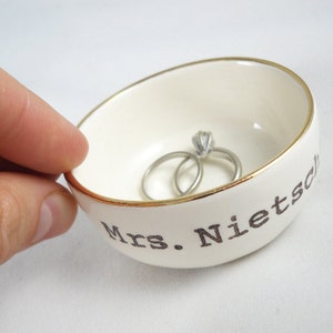 Puede incluir: Plato de anillos de cer&aacute;mica blanca con borde dorado y el texto "Mrs. Nietzsche" en letras negras. Dos anillos est&aacute;n dentro del plato.