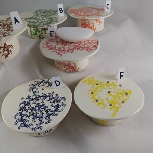May include: Six ceramic pedestals with different colored floral designs. The pedestals are labeled A through F. The pedestals are all white with a raised rim. The designs are in red, green, blue, yellow, and brown.