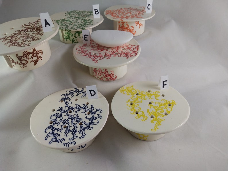 May include: Six ceramic pedestals with different colored floral designs. The pedestals are labeled A through F. The pedestals are all white with a raised rim. The designs are in red, green, blue, yellow, and brown.