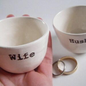 Puede incluir: Dos cuencos de cer&aacute;mica blanca con las palabras "Wife" y "Husband" escritas en ellos. Los cuencos est&aacute;n sobre una superficie blanca con dos anillos dorados delante de ellos.