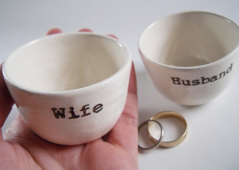 Puede incluir: Dos cuencos de cer&aacute;mica blanca con las palabras "Wife" y "Husband" escritas en ellos. Los cuencos est&aacute;n sobre una superficie blanca con dos anillos dorados delante de ellos.