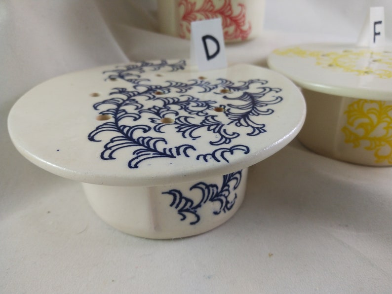 May include: A white ceramic dish with a blue floral design and a matching base. The dish has a letter "D" on a white card in front of it.