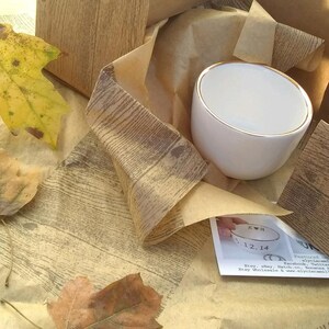 May include: A white ceramic mug with a gold rim. The mug is wrapped in brown paper with a wood grain pattern. The mug has the initials "KH" and the date "12.14" written on it.