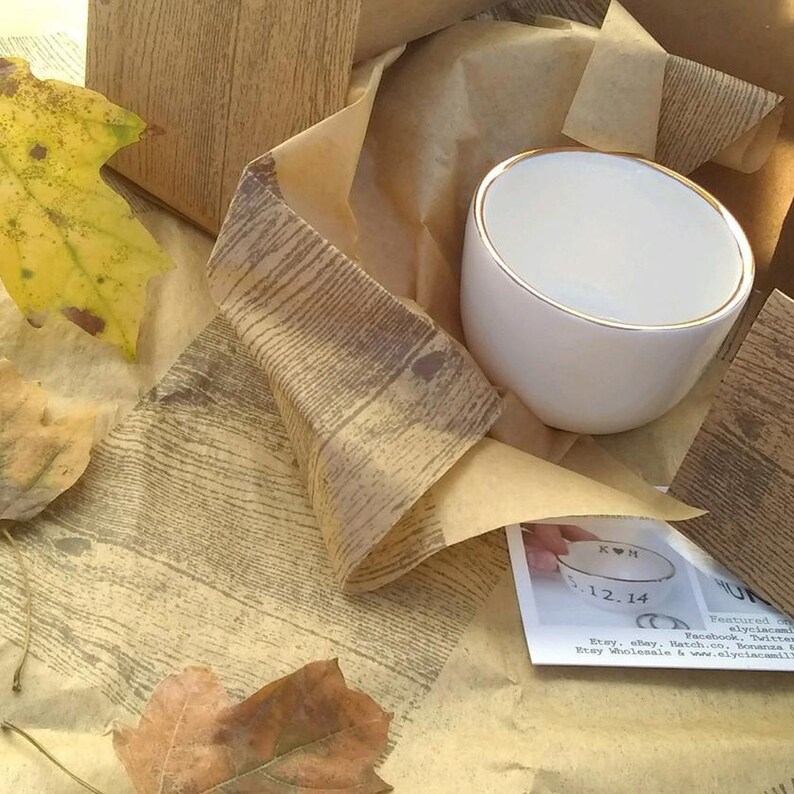 May include: A white ceramic mug with a gold rim. The mug is wrapped in brown paper with a wood grain pattern. The mug has the initials "KH" and the date "12.14" written on it.