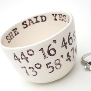 May include: Cream-colored ceramic bowl with the text "SHE SAID YES!" and coordinates in brown. A silver ring with a clear stone is next to the bowl. The bowl is tilted, showing the inside.