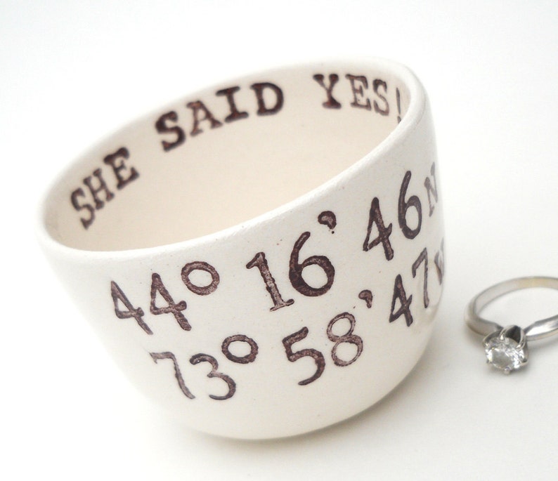 May include: Cream-colored ceramic bowl with the text "SHE SAID YES!" and coordinates in brown. A silver ring with a clear stone is next to the bowl. The bowl is tilted, showing the inside.