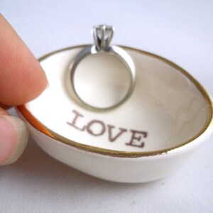 May include: White ceramic ring dish with a gold rim and the word "LOVE" printed in brown. A silver ring with a single diamond is sitting in the dish.
