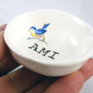 May include: White ceramic trinket dish with a bluebird design and the word "AMI" printed in black.