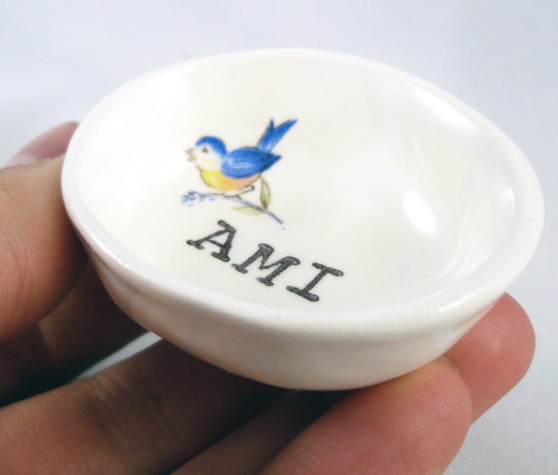 May include: White ceramic trinket dish with a bluebird design and the word "AMI" printed in black.