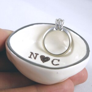 May include: White ceramic ring dish with a silver rim. The dish has the initials "N & C" with a heart in between. A silver engagement ring with a single diamond is sitting in the dish.