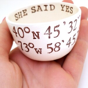 May include: A small, white ceramic bowl with the words "SHE SAID YES" and coordinates printed in brown. The bowl is held in a hand, showcasing its size and design. The coordinates are 40&deg;N 45'32" and 73&deg;W 58'45".