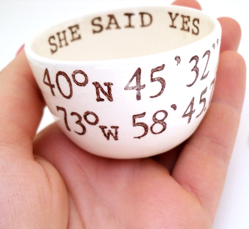 May include: A small, white ceramic bowl with the words "SHE SAID YES" and coordinates printed in brown. The bowl is held in a hand, showcasing its size and design. The coordinates are 40&deg;N 45'32" and 73&deg;W 58'45".