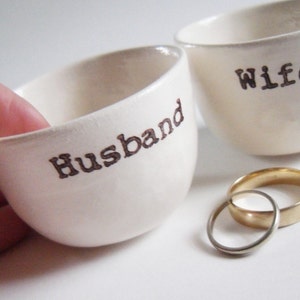 Puede incluir: Dos cuencos de cer&aacute;mica blanca con las palabras "Husband" y "Wife" inscritas en ellos. Dos alianzas de oro se colocan delante de los cuencos.