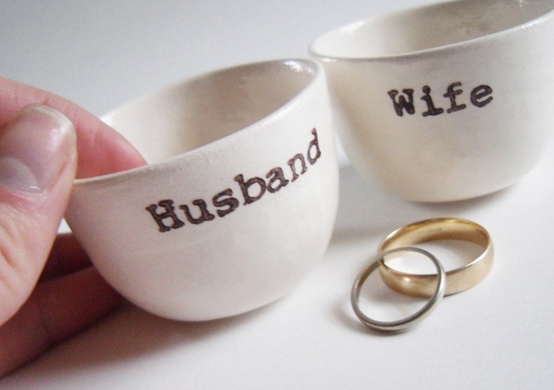 Puede incluir: Dos cuencos de cer&aacute;mica blanca con las palabras "Husband" y "Wife" inscritas en ellos. Dos alianzas de oro se colocan delante de los cuencos.