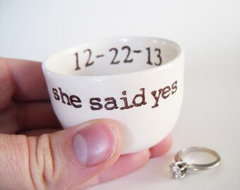May include: White ceramic ring dish with the text "12-22-13 she said yes" in brown lettering. A silver engagement ring is sitting next to the dish.