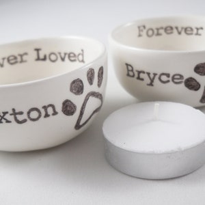 May include: Two white ceramic bowls with black paw prints and the text "Forever Loved" and the names "Braxton" and "Bryce". A white tea light candle is in the foreground.
