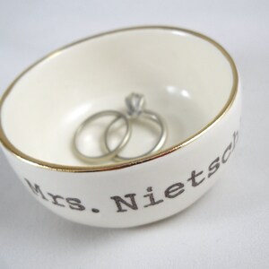 Puede incluir: Plato de cer&aacute;mica blanco para anillos con borde dorado y el texto "Mrs. Nietzsche" en letras negras. Dos anillos de plata est&aacute;n dentro del plato.