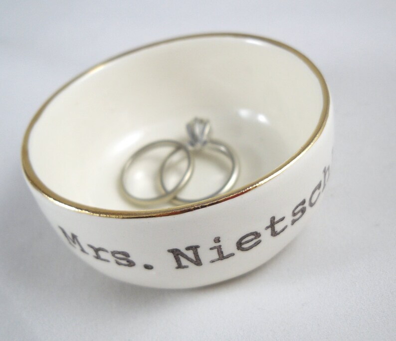 Puede incluir: Plato de cer&aacute;mica blanco para anillos con borde dorado y el texto "Mrs. Nietzsche" en letras negras. Dos anillos de plata est&aacute;n dentro del plato.