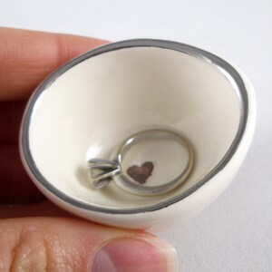 May include: A white ceramic ring dish with a silver rim. The dish has a small heart design in the center and a silver ring is resting inside.