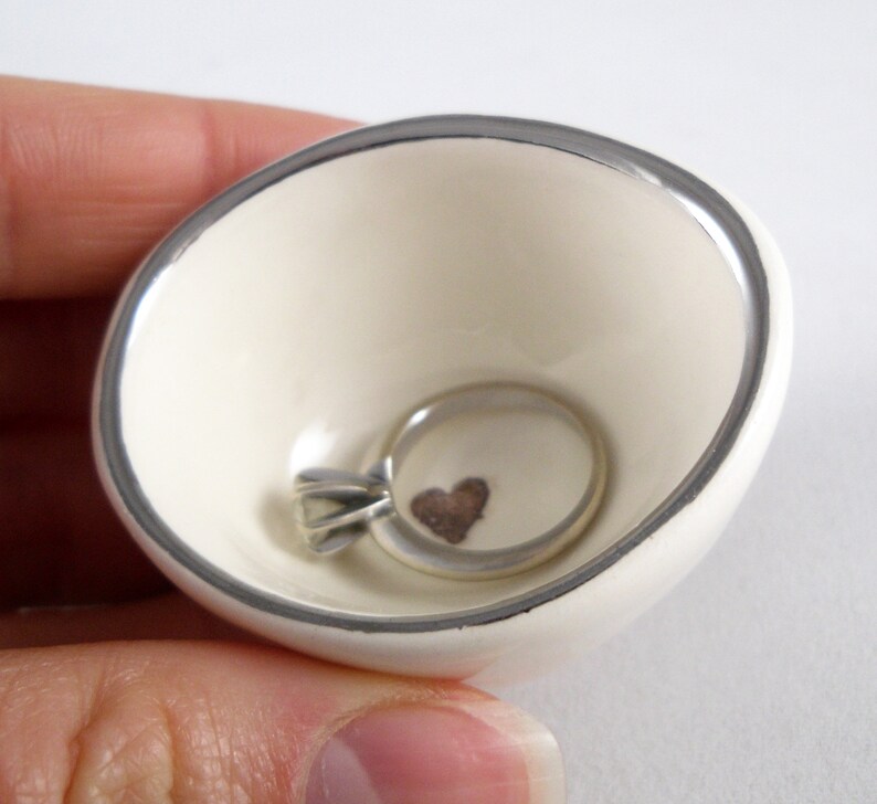 May include: A white ceramic ring dish with a silver rim. The dish has a small heart design in the center and a silver ring is resting inside.