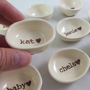 May include: A set of small, white ceramic bowls with brown lettering and hearts. The bowls are labelled with the names "kat", "jamie", "chelse", "baby", and other names.